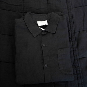 May include: A black linen shirt with a button-up front and a single pocket on the left side. The shirt is folded and placed on a black quilted fabric.