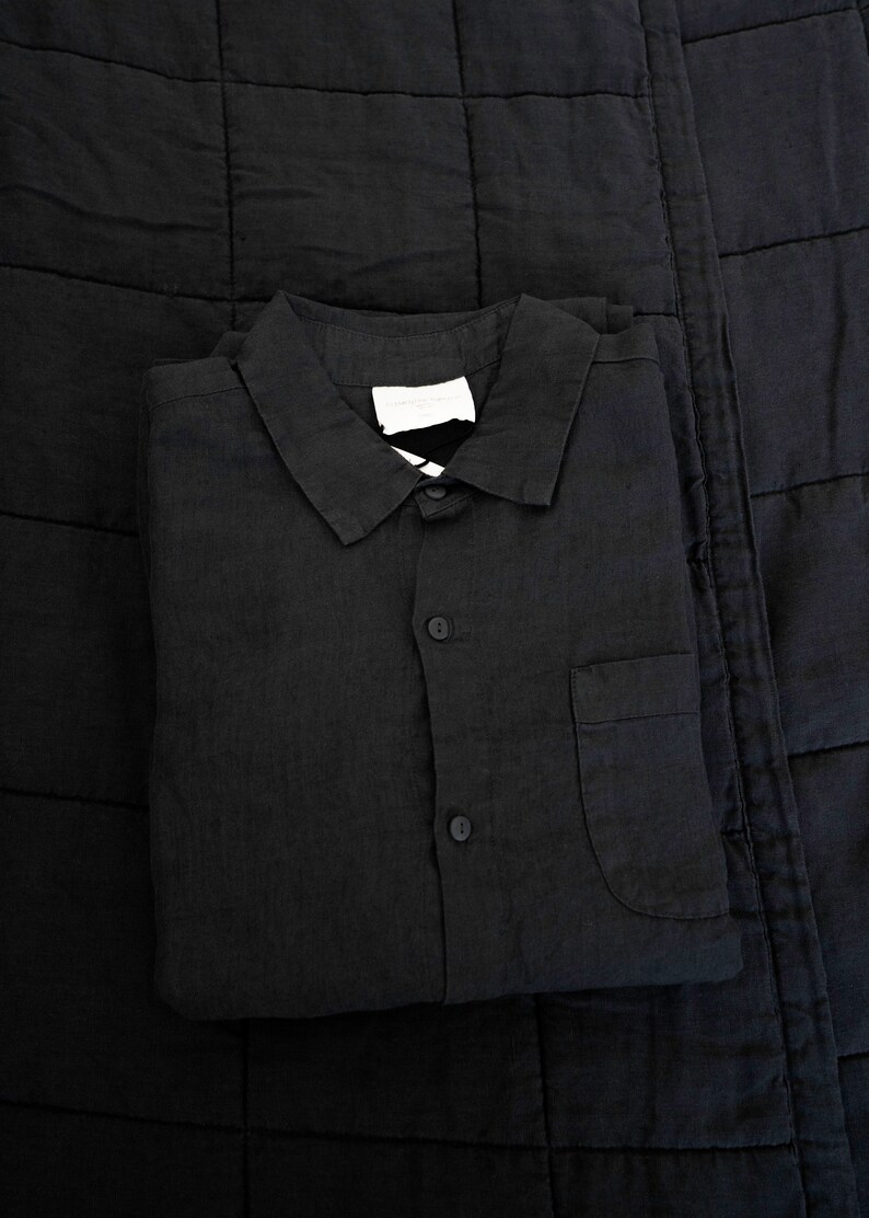 May include: A black linen shirt with a button-up front and a single pocket on the left side. The shirt is folded and placed on a black quilted fabric.