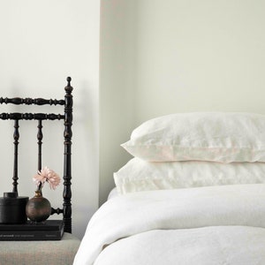 Puede incluir: Una cama blanca con un edred&oacute;n de lino blanco y dos almohadas de lino blanco. Una silla de madera negra con un arreglo floral en el asiento est&aacute; al lado de la cama.