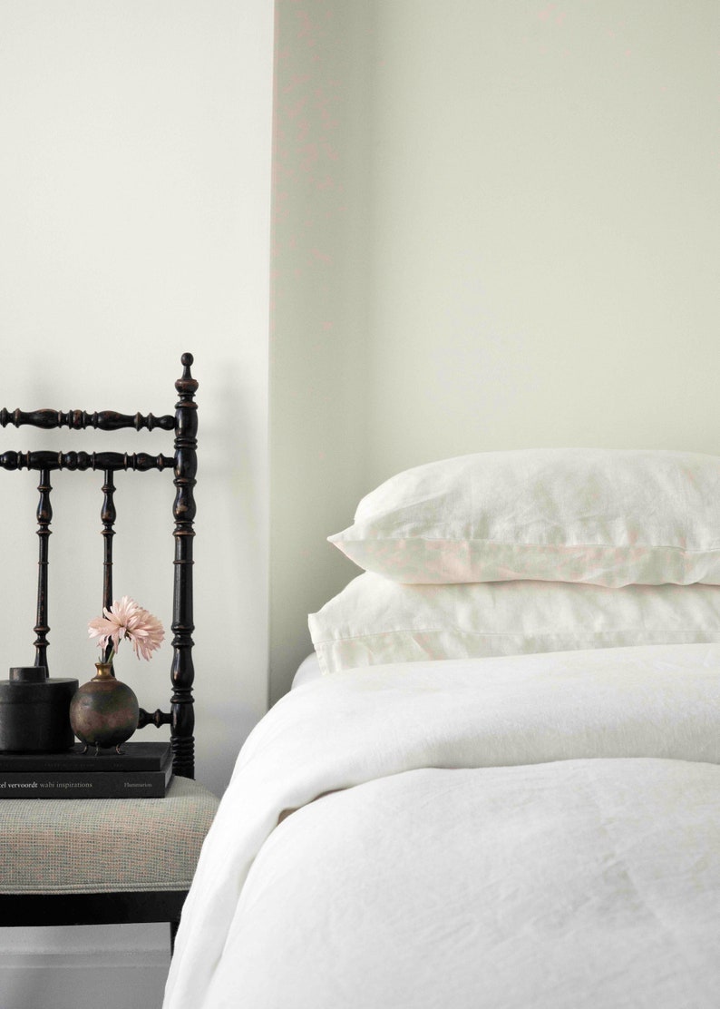 Puede incluir: Una cama blanca con un edred&oacute;n de lino blanco y dos almohadas de lino blanco. Una silla de madera negra con un arreglo floral en el asiento est&aacute; al lado de la cama.