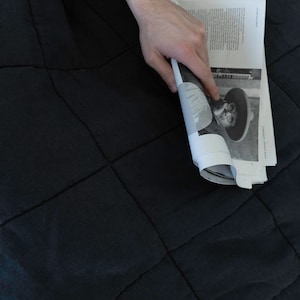May include: A person is holding a magazine with a black and white photo of a person wearing a hat and sunglasses. The magazine is on a black quilted blanket.