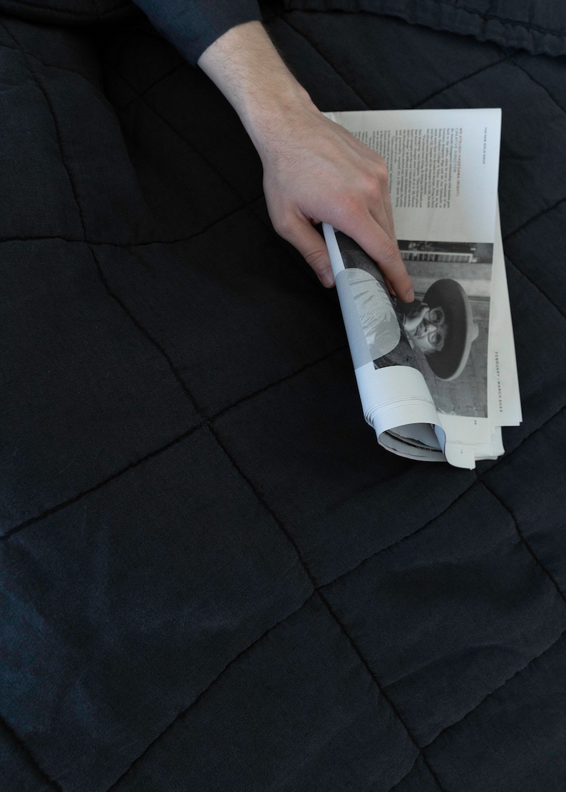 May include: A person is holding a magazine with a black and white photo of a person wearing a hat and sunglasses. The magazine is on a black quilted blanket.