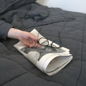 May include: A grey quilted bedspread with a folded newspaper and a pair of black spectacles on top.