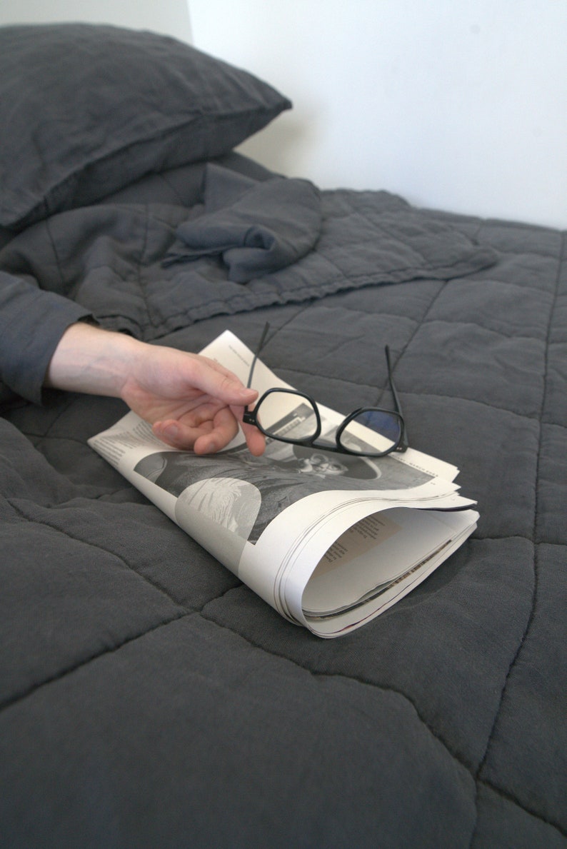 May include: A grey quilted bedspread with a folded newspaper and a pair of black spectacles on top.