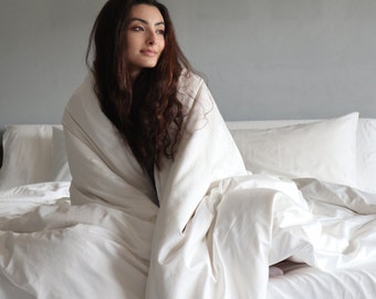 May include: A woman sits in a bed, wrapped in a white duvet, looking off to the side. The bed is made with white sheets and pillows.