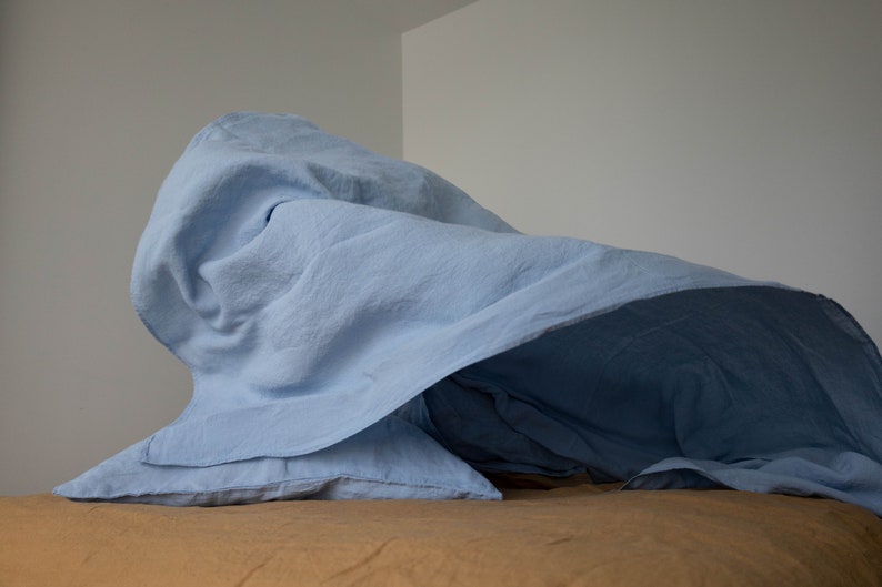 Puede incluir: Una s&aacute;bana de lino azul claro drapeada sobre un cubrecama de lino marr&oacute;n. La s&aacute;bana est&aacute; arrugada en el medio, creando un aspecto suave y acogedor.