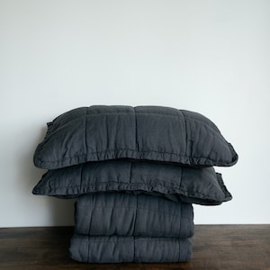 May include: A stack of three dark grey quilted throw cushions. The cushions are made from a soft, textured fabric and have a subtle sheen.