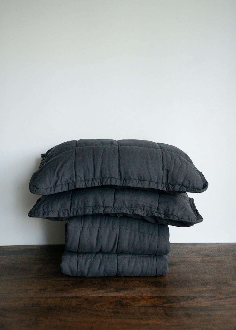 May include: A stack of three dark grey quilted throw cushions. The cushions are made from a soft, textured fabric and have a subtle sheen.