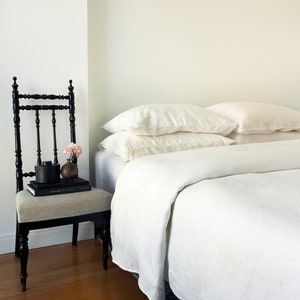 May include: A bedroom scene with a bed dressed in white linen bedding and pillows. A black chair with a patterned seat holds books and decorative items. The room has a minimalist aesthetic with a wooden floor.