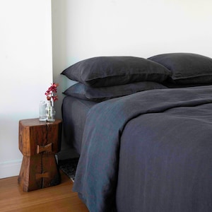 Puede incluir: Una funda n&oacute;rdica y fundas de almohada de lino gris oscuro en una cama con una mesita de noche de madera y un jarr&oacute;n de flores.