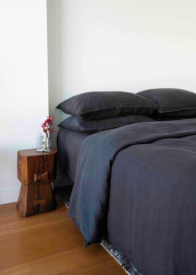 Puede incluir: Una funda n&oacute;rdica y fundas de almohada de lino gris oscuro en una cama con una mesita de noche de madera y un jarr&oacute;n de flores.