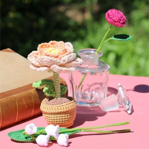 Peut inclure: Un ensemble de fleurs crochetées à la main de différentes couleurs. Une grande fleur pêche et jaune est dans un petit pot marron. Une fleur rose est dans une bouteille en verre transparente. Des fleurs blanches et une feuille verte sont également présentes.
