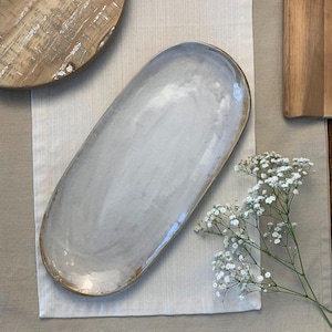 May include: A white oval ceramic serving platter with a slightly textured surface. The platter is on a light brown linen tablecloth.
