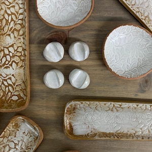 May include: A collection of handmade ceramic serving dishes and small bowls with a leaf pattern. The dishes have a white glaze over the leaf design and a brown rim. There are also four small, white, rounded ceramic pieces.