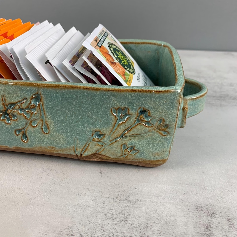 May include: A rectangular, turquoise ceramic tea caddy with floral relief detailing. The caddy has a handle on one side and is filled with tea and sweetener packets. The caddy is on a light gray surface.