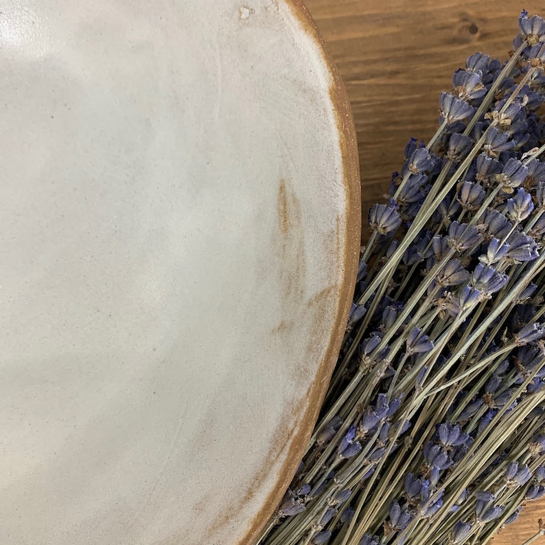 Puede incluir: Un cuenco de cer&aacute;mica blanca con un borde marr&oacute;n. El cuenco est&aacute; vac&iacute;o y se encuentra sobre una superficie de madera. Hay flores de lavanda secas en el fondo.