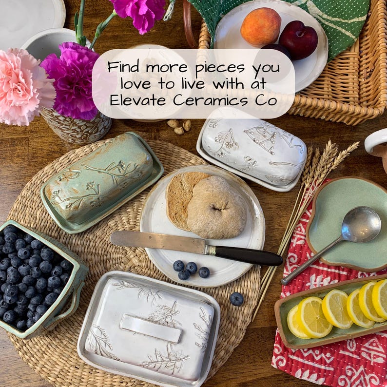 May include: An overhead shot of a table setting featuring ceramic serving dishes, including butter dishes, a square bowl of blueberries, and plates with food. The text "Find more pieces you love to live with at Elevate Ceramics Co" is visible.