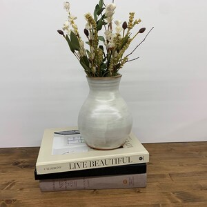 May include: A white ceramic vase with a rounded body and a slightly flared opening, filled with a bouquet of dried white flowers and brown stems. The vase is sitting on top of two books, one with the title "Live Beautiful" and the other with the title "Art Through the Ages".