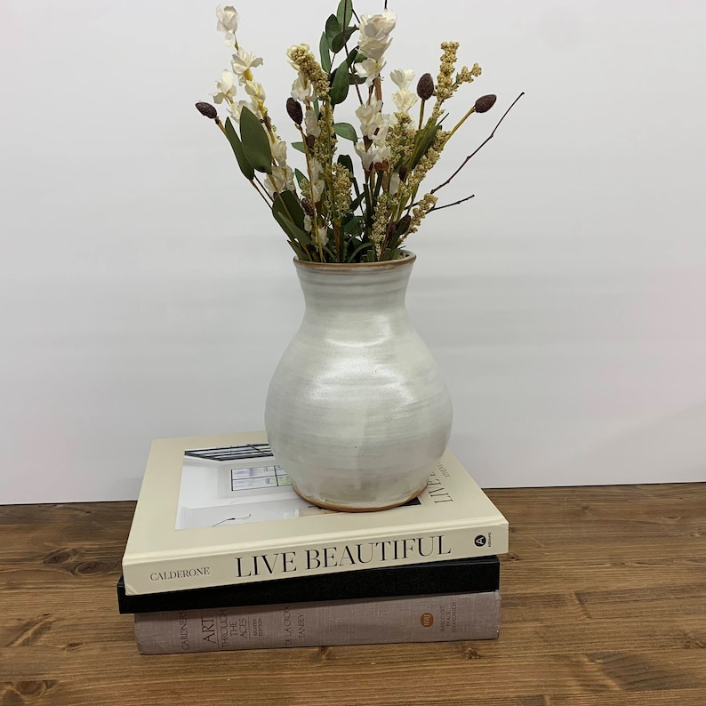 May include: A white ceramic vase with a rounded body and a slightly flared opening, filled with a bouquet of dried white flowers and brown stems. The vase is sitting on top of two books, one with the title "Live Beautiful" and the other with the title "Art Through the Ages".