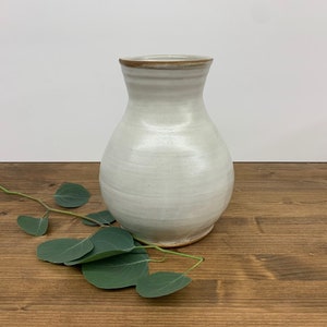 May include: A white ceramic vase with a brown rim. The vase has a rounded body and a narrow neck. It is sitting on a wooden surface with a green leafy branch.