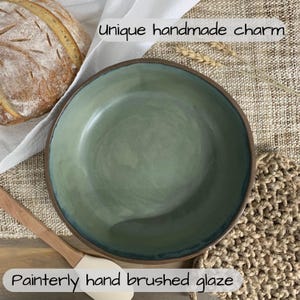 May include: Handmade ceramic bowl with a painterly, hand-brushed glaze in shades of green and brown. The bowl has a unique, rustic charm. The rim is a darker brown, and the interior has a smooth, glossy finish. The text in the image says "Unique handmade charm" and "Painterly hand brushed glaze."
