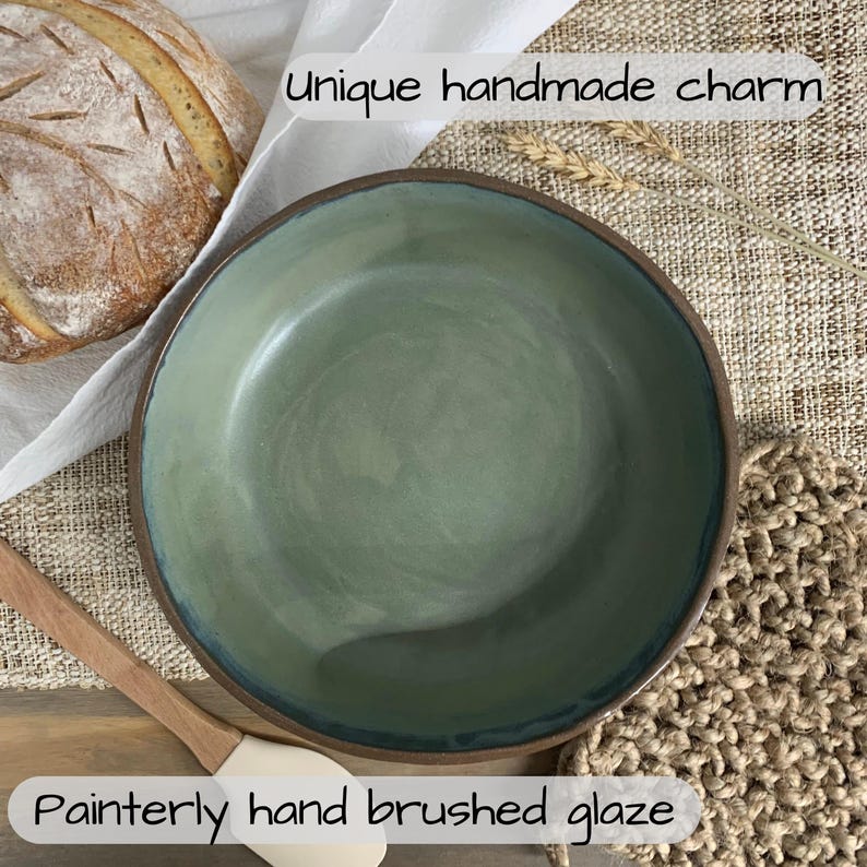 May include: Handmade ceramic bowl with a painterly, hand-brushed glaze in shades of green and brown. The bowl has a unique, rustic charm. The rim is a darker brown, and the interior has a smooth, glossy finish. The text in the image says "Unique handmade charm" and "Painterly hand brushed glaze."