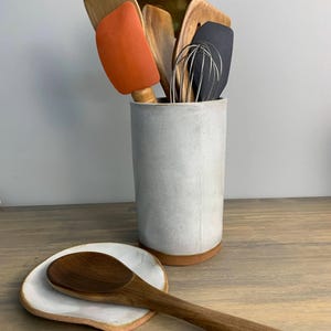 May include: A collection of kitchen utensils in a white ceramic utensil holder. The utensils include wooden spoons, spatulas, and a whisk. A wooden spoon rests on a small, white, ceramic spoon rest.