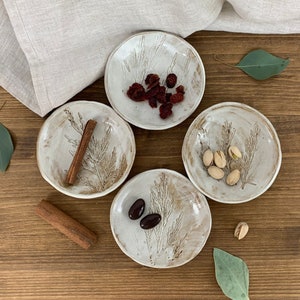 May include: Four small white ceramic bowls with a brown leaf design. Each bowl contains a different snack: cranberries, cinnamon sticks, pistachios, and chocolate candies.