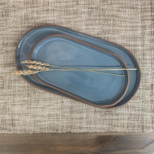 May include: Set of three oval-shaped ceramic serving platters in varying sizes. The platters are a muted blue with a brown rim. Wheat stalks are placed on the largest platter, which is on a woven beige surface.