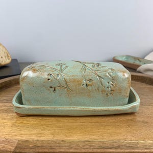 May include: A rectangular, ceramic butter dish in a light teal color with a floral design on the lid. The dish sits on a matching tray, and the overall aesthetic is rustic and handmade. The butter dish is a kitchenware item.