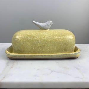 May include: A ceramic butter dish with a yellow speckled lid and a matching base. The lid is topped with a small, white bird figurine. The dish is designed to hold a standard size of butter.