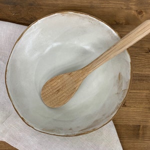 Puede incluir: Un bol de cer&aacute;mica blanco con una superficie ligeramente texturizada y una cuchara de madera en su interior. El bol tiene un borde marr&oacute;n sutil.