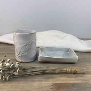 May include: A set of two white ceramic pieces with a textured surface. The set includes a small square dish and a taller cylindrical cup.
