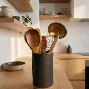 May include: A black ceramic utensil holder with a terracotta base holds wooden spoons, a whisk, and other kitchen tools. The holder sits on a light wood countertop, with wooden shelves and kitchenware in the background. A brass cooking utensil is also visible.