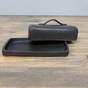 May include: Black ceramic butter dish with a lid and a handle. The dish is on a wooden surface.