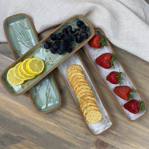 May include: A collection of rectangular ceramic serving platters. Two platters are a light teal color with a leaf design and contain sliced lemons and grapes. Two other platters are white with a leaf design and contain crackers and strawberries.