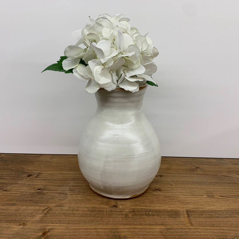 May include: White ceramic vase with a rounded body and a slightly flared opening, holding a bouquet of white hydrangeas.