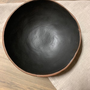 May include: A large, black ceramic bowl with a brown rim. The bowl is empty and sitting on a light brown surface.