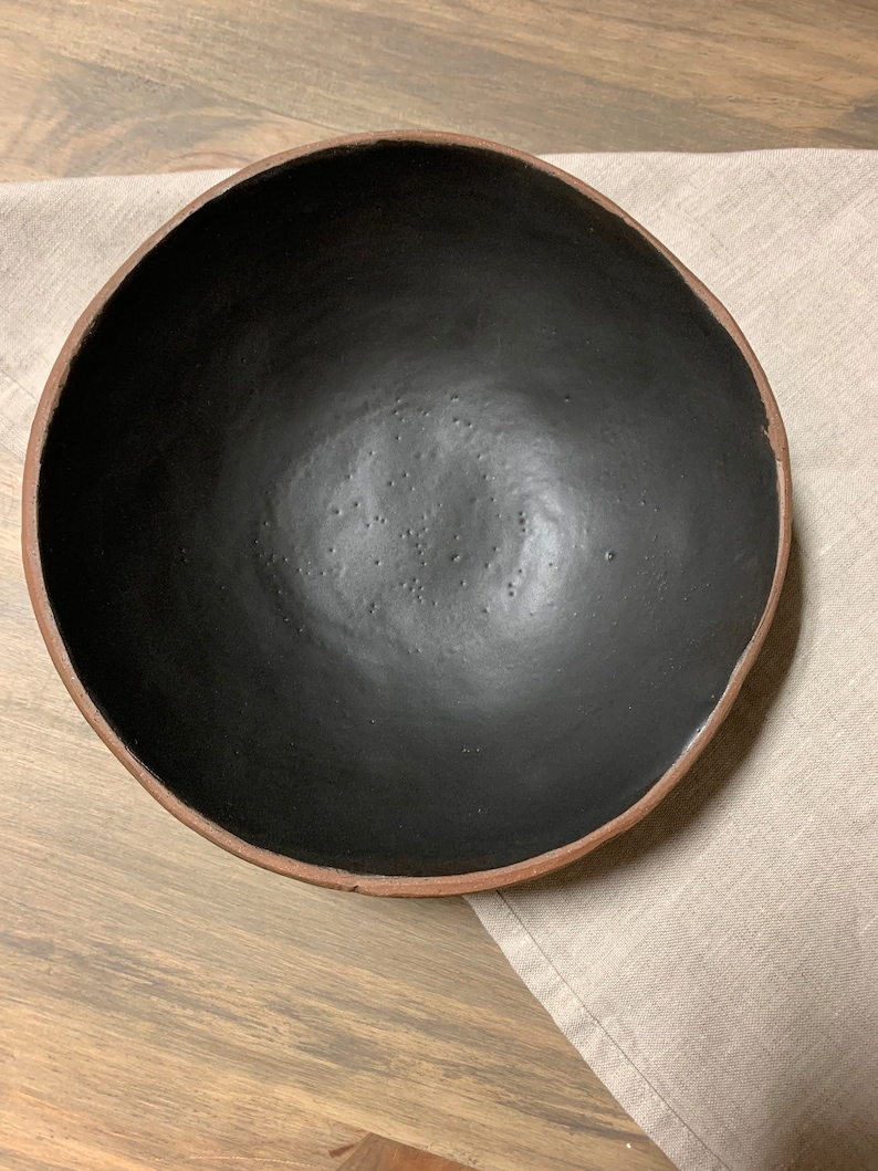 May include: A large, black ceramic bowl with a brown rim. The bowl is empty and sitting on a light brown surface.