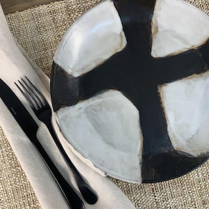 May include: A modern, abstract ceramic plate with a black and white design. The plate is paired with a black fork and knife, resting on a beige linen napkin and a woven placemat. This set is perfect for a contemporary dining experience.