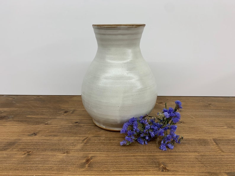 May include: A white ceramic vase with a rounded body and a slightly flared opening. The vase is sitting on a wooden surface with purple flowers in front of it.