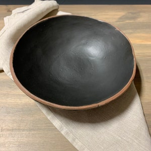 May include: A large, black ceramic bowl with a brown rim. The bowl is sitting on a beige linen napkin.