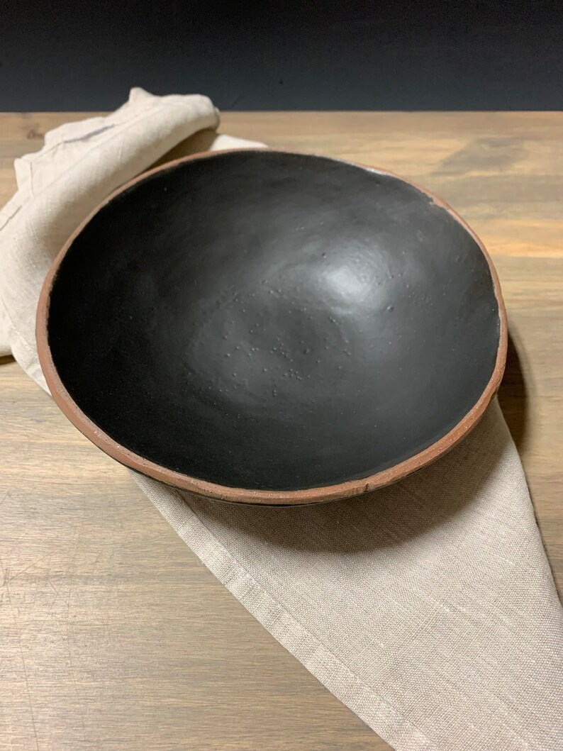 May include: A large, black ceramic bowl with a brown rim. The bowl is sitting on a beige linen napkin.