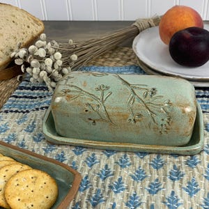 May include: A rectangular, turquoise ceramic butter dish with a lid, featuring an embossed floral design. The dish sits on a matching base. The background includes a blue and white patterned cloth, crackers, and fruit.