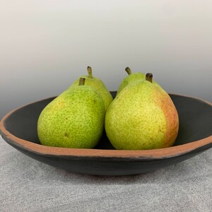 May include: Four green pears in a black ceramic bowl with a brown rim. The bowl is sitting on a gray fabric surface.