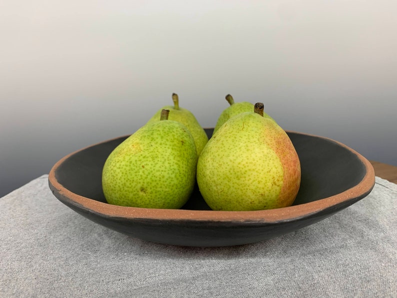 May include: Four green pears in a black ceramic bowl with a brown rim. The bowl is sitting on a gray fabric surface.