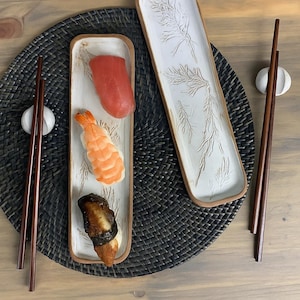 May include: Sushi serving set featuring two rectangular ceramic plates with a white glaze and a brown rim. One plate holds sushi: tuna, shrimp, and eel. The other plate has a botanical design. Chopsticks and chopstick rests complete the set.