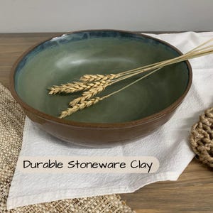 May include: A handmade stoneware bowl with a green interior and a brown exterior. The rim has a blue-green glaze. The bowl is filled with wheat stalks and rests on a white cloth. The text "Durable Stoneware Clay" is visible.