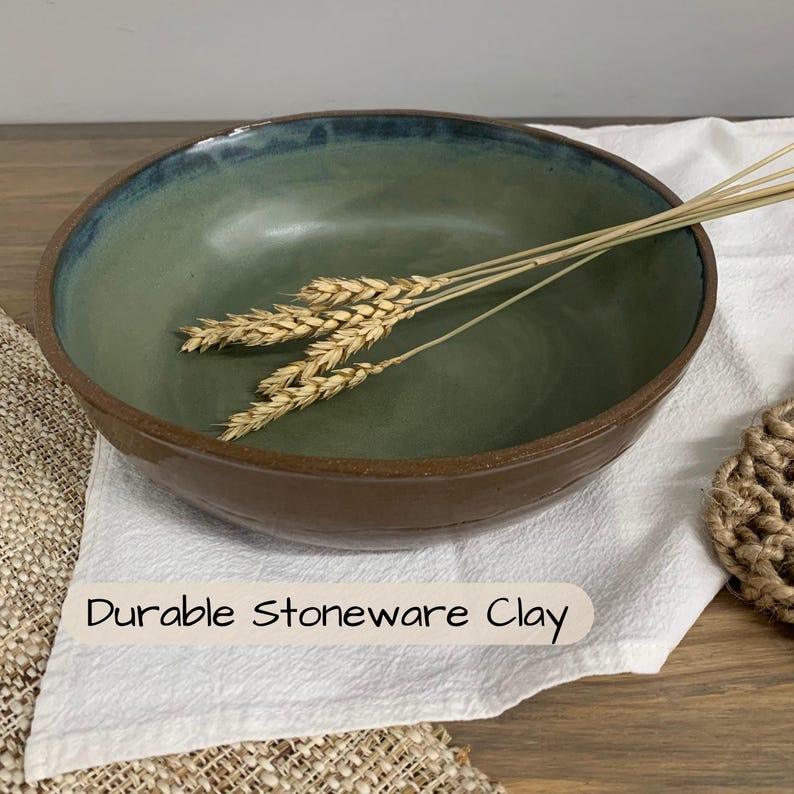 May include: A handmade stoneware bowl with a green interior and a brown exterior. The rim has a blue-green glaze. The bowl is filled with wheat stalks and rests on a white cloth. The text "Durable Stoneware Clay" is visible.