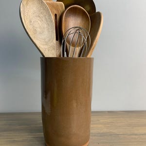 May include: A collection of wooden kitchen utensils, including spoons, spatulas, and a whisk, are displayed in a brown ceramic utensil holder. The holder is cylindrical and sits on a wooden surface.